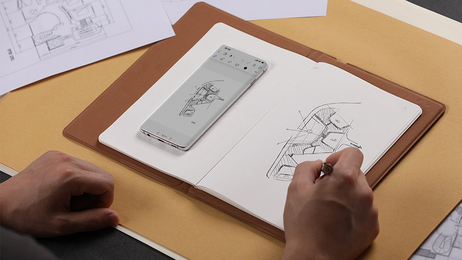 A person uses a stylus to draw a floor plan in a notebook, while a smartphone displaying a similar digital sketch rests on the open pages nearby.