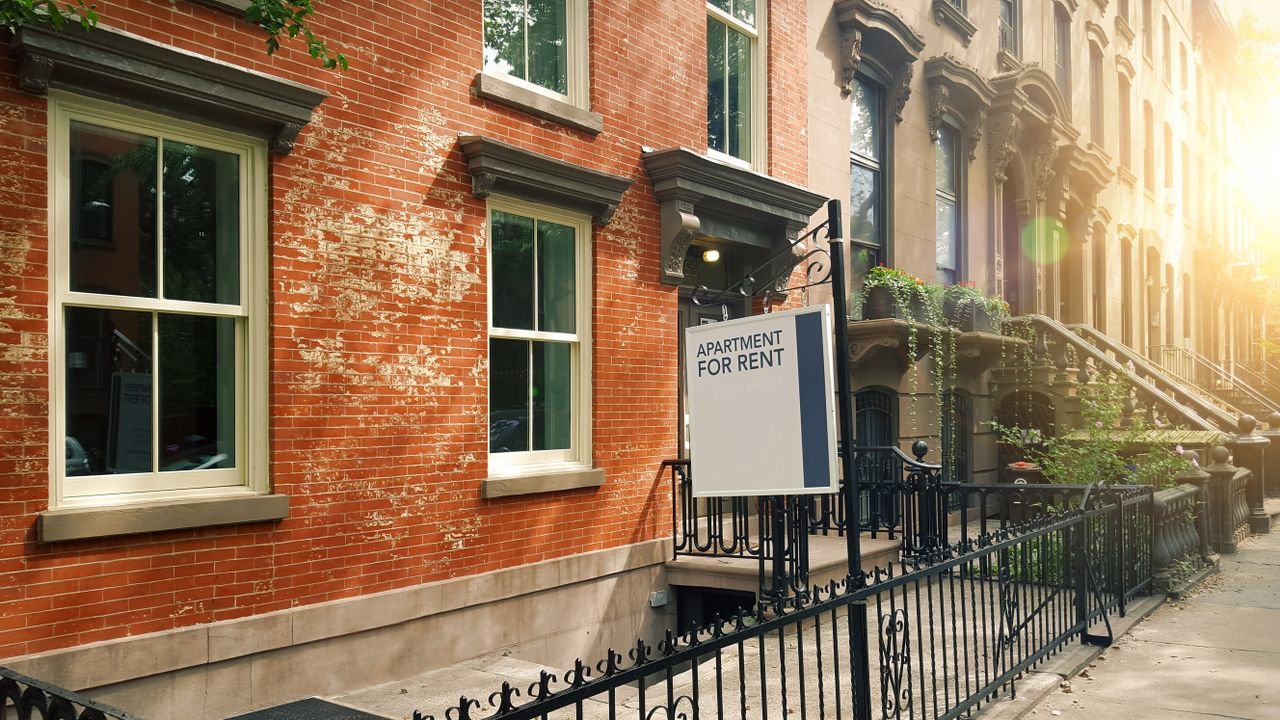 The outside of a traditional Brooklyn, New York, apartment building taken from the sidewalk at sunset. There is a white and blue sign hanging outside, which says &#039;Apartment for rent&#039;