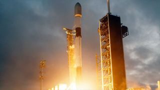a white rocket with black stripes lifts off its launch pad under an overcast sky