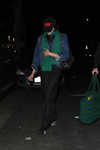 Kaia Gerber wears a baseball cap, green scarf, denim jacket, black pants, and black and white Onitsuka Tiger sneakers.