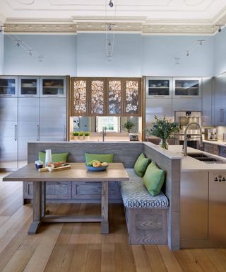 A kitchen with a gray wall and cabinets, a dark gray wooden L-shaped kitchen island with white surfaces, and a corner seat attached to it with green throw pillows and a table