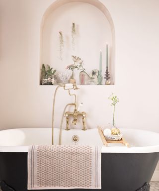 A small bathroom with a beige wall with an arched shelf with candles and flowers, a curved black bath tub with gold faucets and a wooden bath tray, and a beige bath mat hanging on the side of it