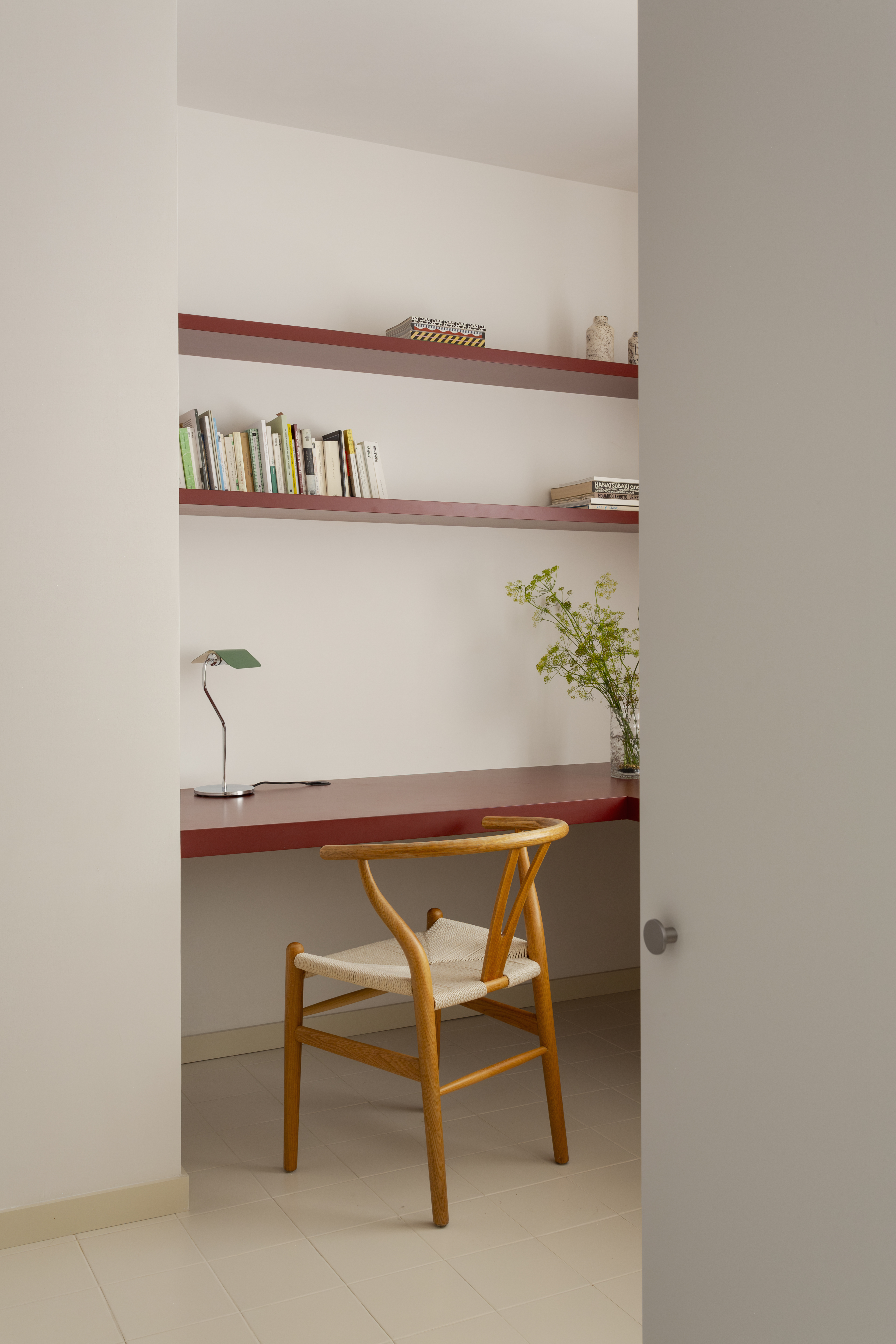 A home office with built in long red desk and red matching red shelves above with some books and decorative pieces with a wooden chair and a desk lamp and vase filled with foliage