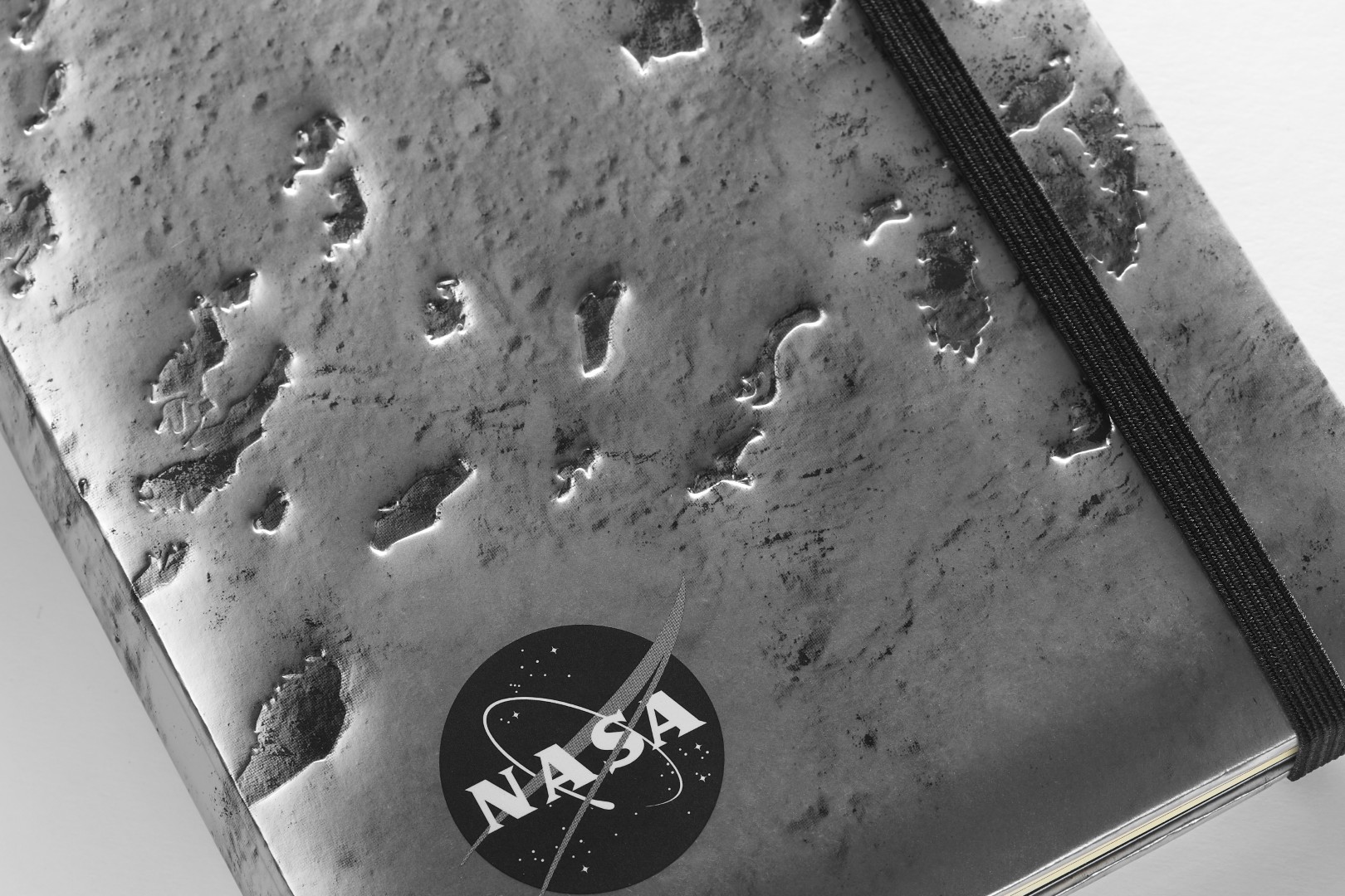 Close up a NASA-inspired Limited Edition Notebook from Moleskine in dark silver. The cover is a close up of the moon's surface. The notebook has a black elastic closure. A black and white version of the NASA logo is in the bottom left corner.