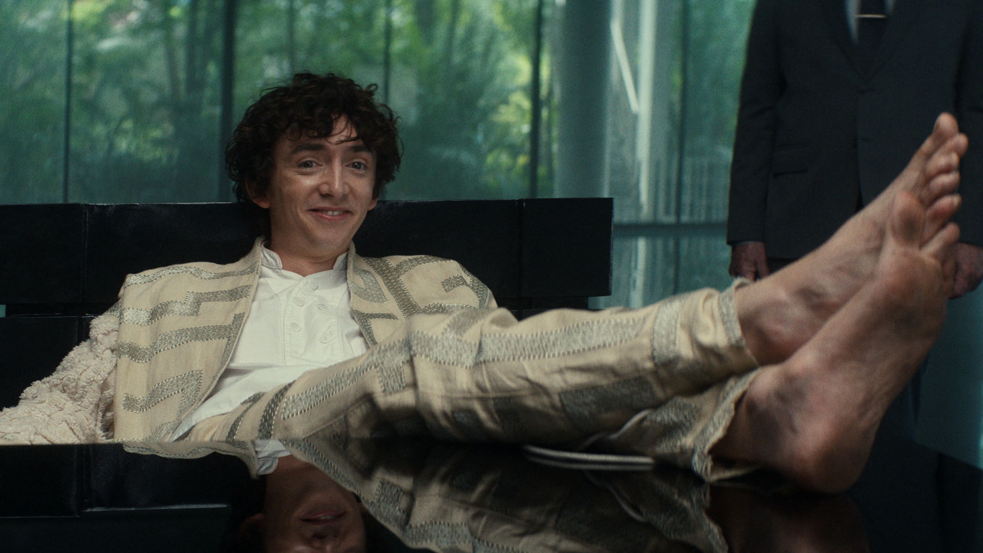Still from the TV show Alien: Earth. A grinning man (wearing a beige and silver suit jacket and pants) is sitting at a desk with his bare feet on the desk.