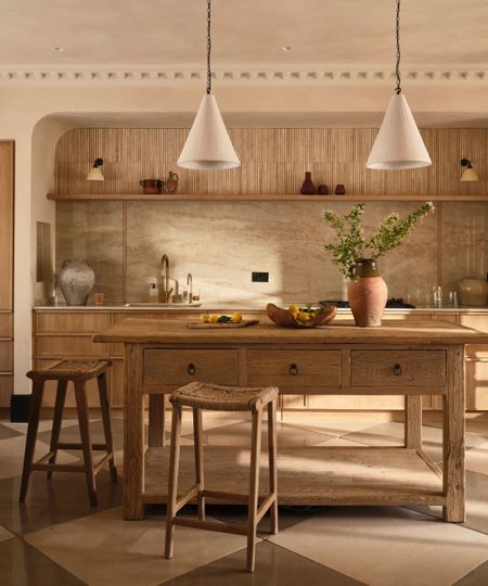 A neutral kitchen with checkered flooring, a large wooden kitchen island and cabinetry, two rattan bar stools, and a marble backsplash. On the island is a large vase of flowers and a bowl of fruit.