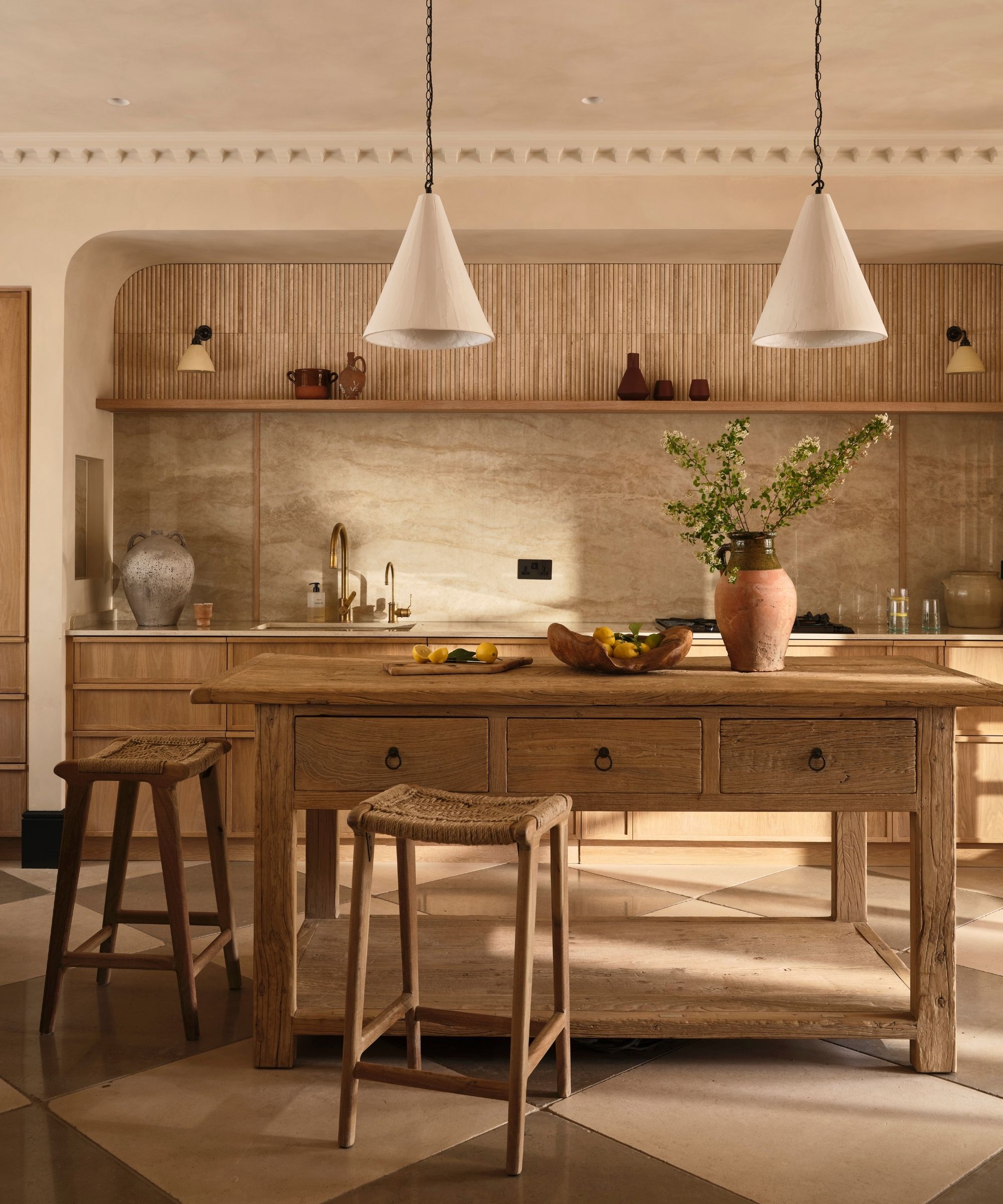 A neutral kitchen with checkered flooring, a large wooden kitchen island and cabinetry, two rattan bar stools, and a marble backsplash. On the island is a large vase of flowers and a bowl of fruit.