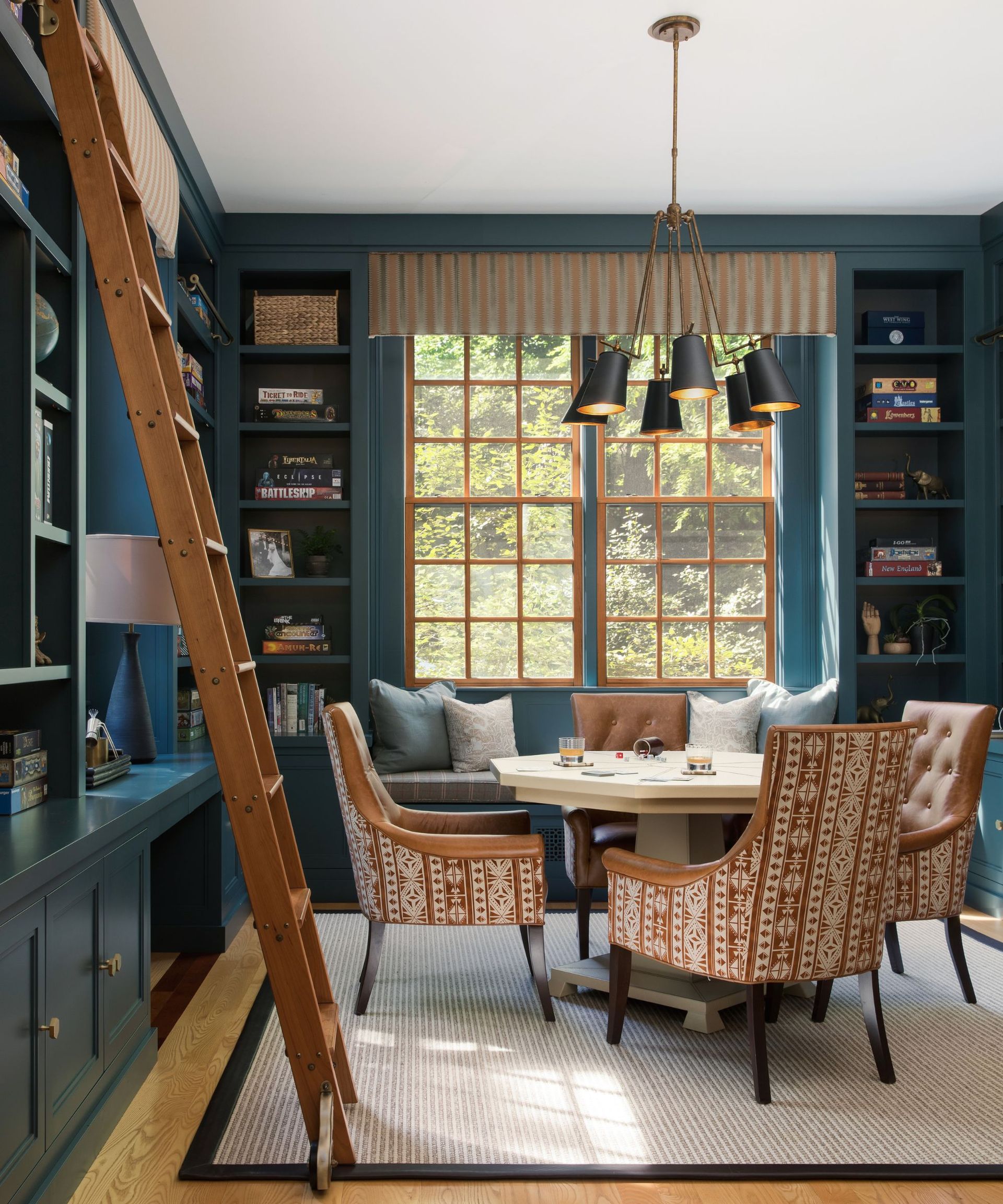 Games room with blue walls and built-in bookshelves, a large window, a wooden ladder, a games table with armchairs around it, and a built-in bench with blue and