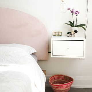 Floating nightstand with drawer in white styled with potted orchid and trinkets, next to curved headboard in pale pink, and exposed bulb pendant suspended above.