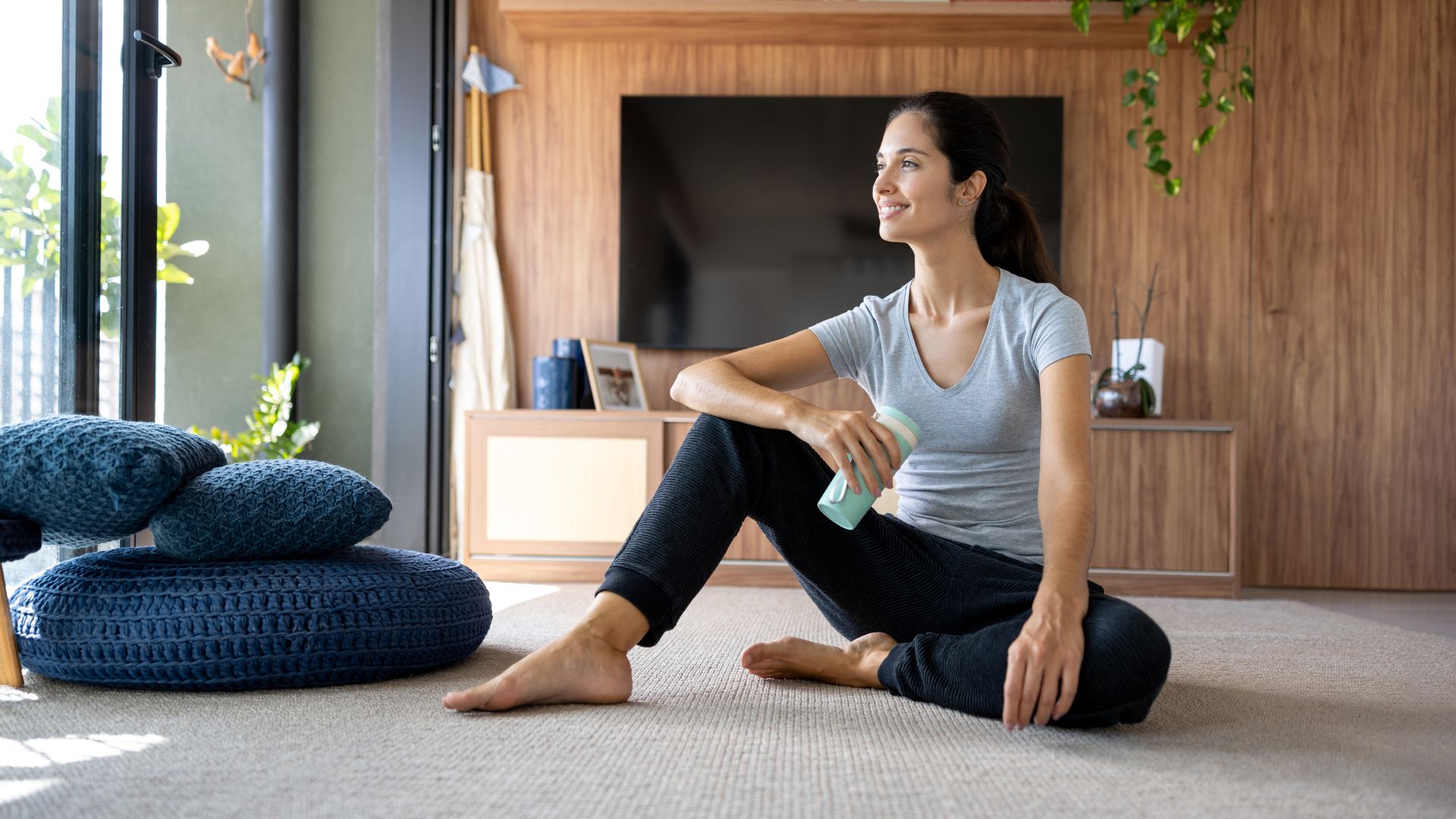 Woman sitting down on carpet with leg crossed, looking out of window, sofa and television behind her, setting up to do the clamshell exercise