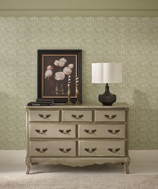 room with green wallpaper, pale green antiqued chest of drawers with table lamp on top with a white curved shade