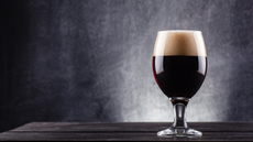 A glass of dark beer on a wooden surface with a black background.