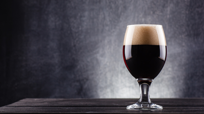 A glass of dark beer on a wooden surface with a black background.