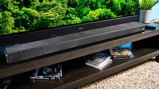 The Polk Audio Signa S4 being used in a lounge with a large TV. On the TV screen is a green forest scene. In the background is a blue wall.