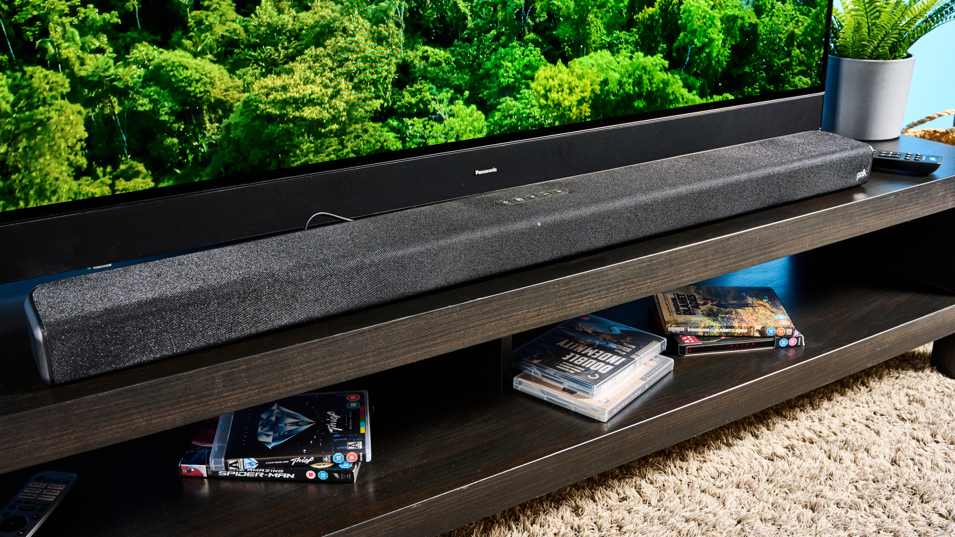 The Polk Audio Signa S4 being used in a lounge with a large TV. On the TV screen is a green forest scene. In the background is a blue wall.