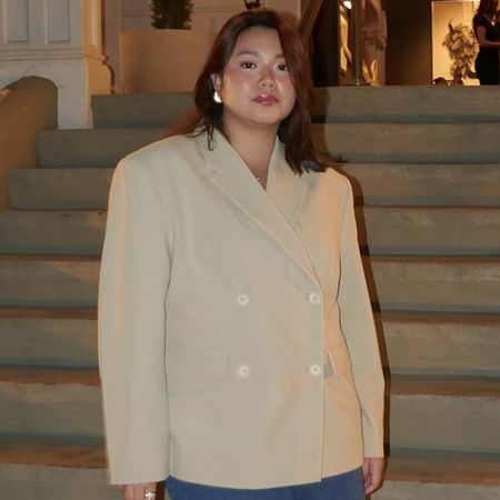 Woman wearing oat-colored blazer with gold earrings, standing in front of staircase.