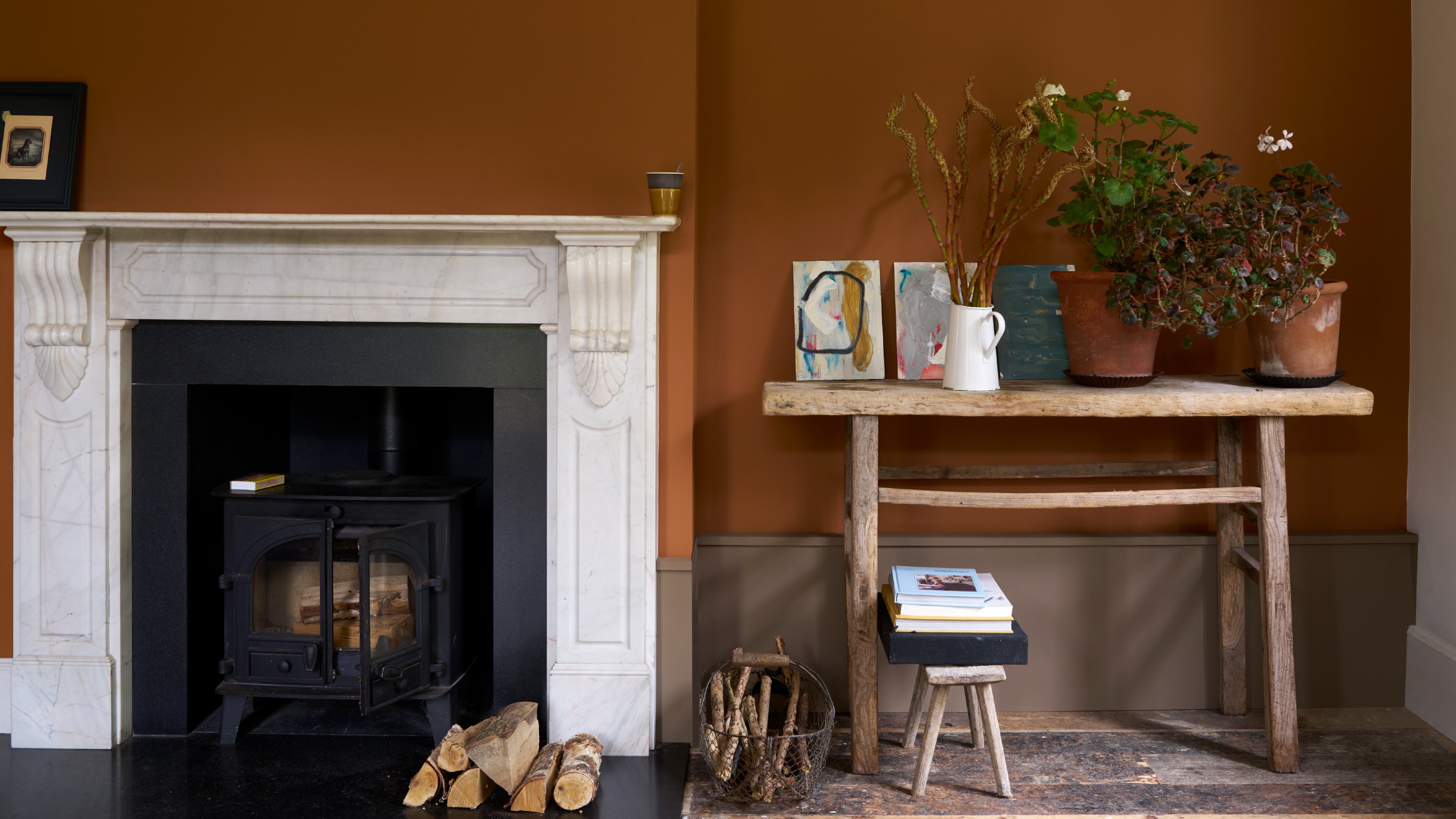 A wall painted in Farrow &amp; Ball&#039;s Marmelo shade with a marble fireplace and a rustic wooden console table displaying potted plants and artwork