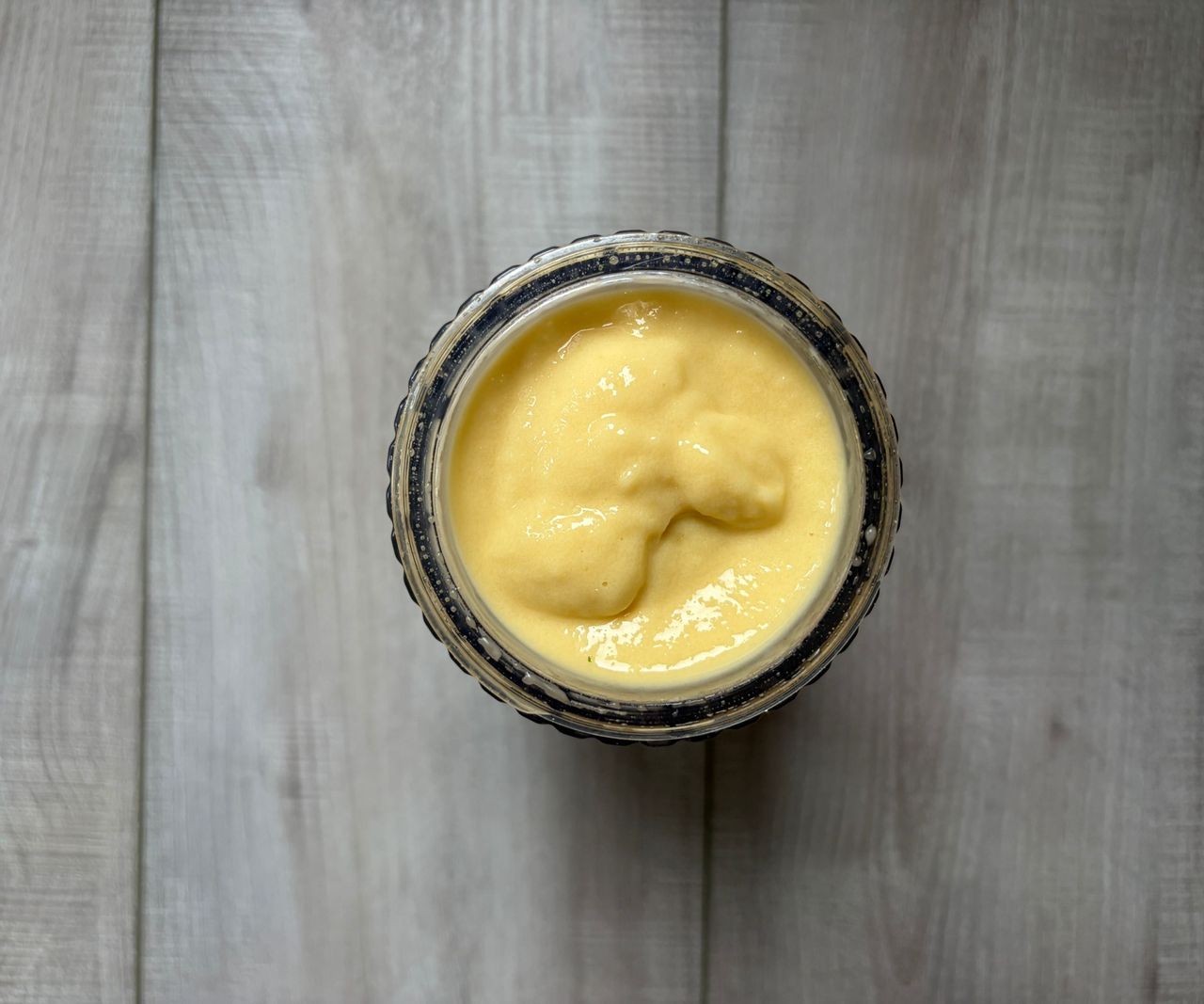A top down view of a yellow smoothie inside a small blender pitcher, on a wooden table.