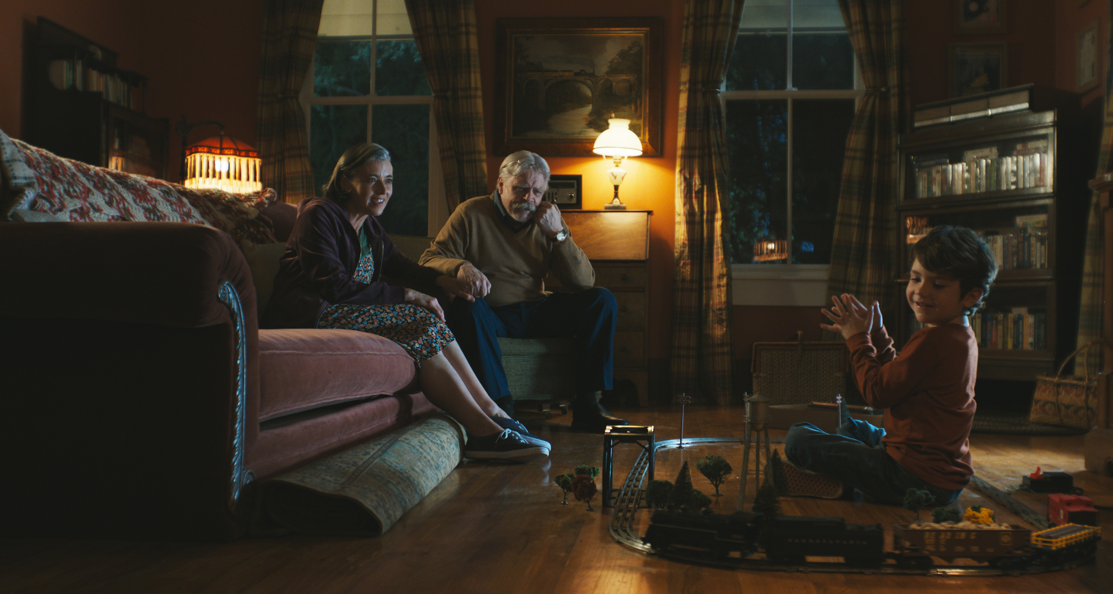 A couple (Mia Sara as Sarah Krantz, Mark Hamill as Albie Krantz) sits on a living room couch and watches a child (Cody Flanagan as Chuck Krantz) play with a model train, in 'The Life of Chuck.'