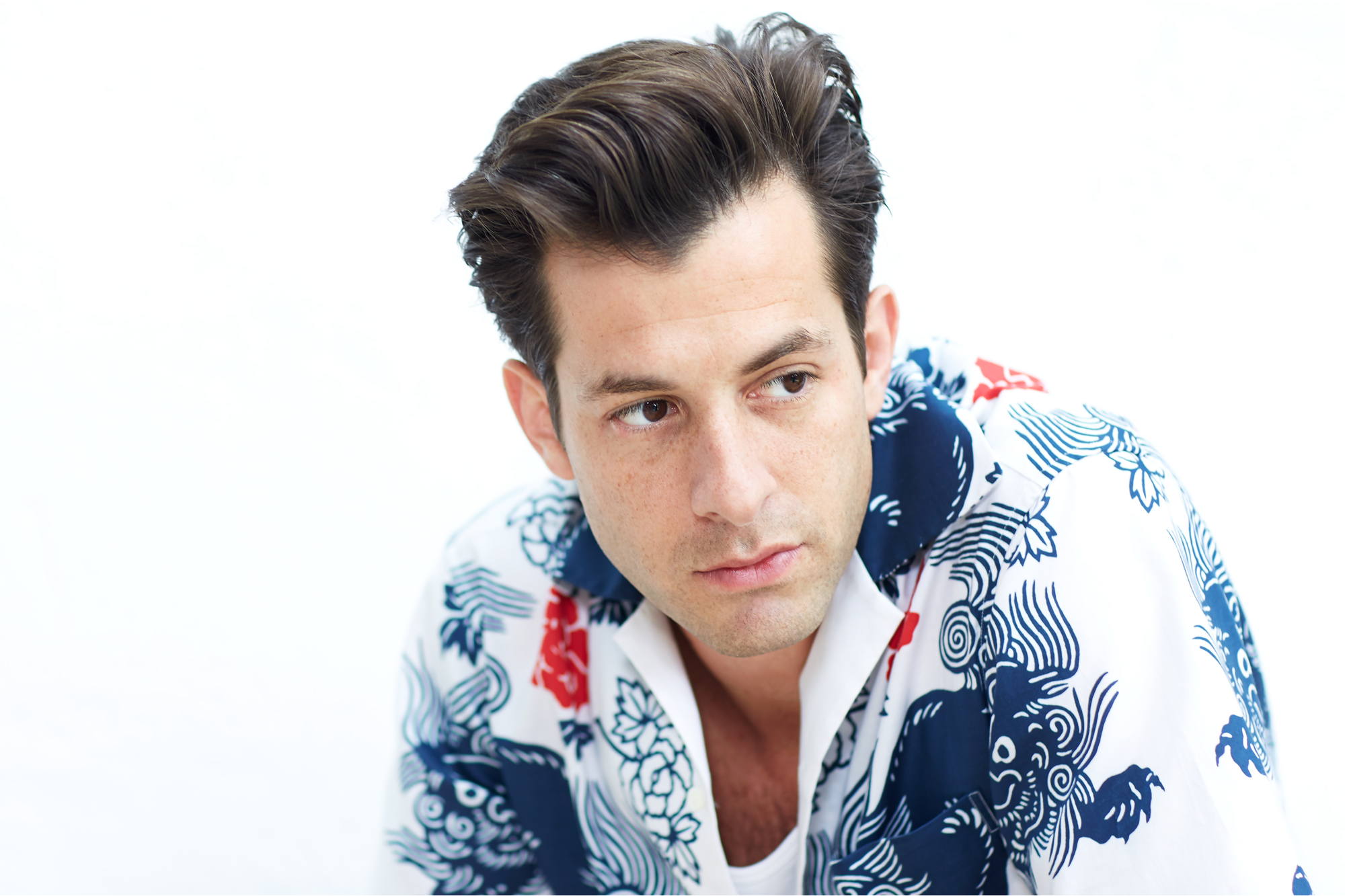 A close-up portrait of Mark Ronson with dark hair, looking off-camera while wearing a white shirt with a bold blue and red patterned print.