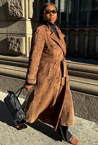 The winter capsule wardrobe item, cozy sneakers, is shown in a photo of a woman walking down the sidewalk wearing black sunglasses, a black turtleneck, a brown suede trench coat, black trousers, brown suede sneakers, and a black shoulder bag