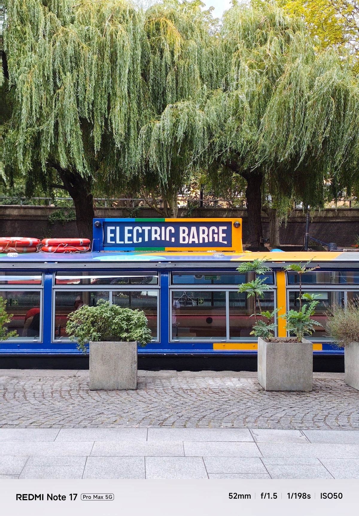 Colorful Electric Barge moored on a canal beneath large willow trees.