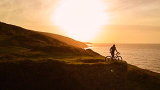 Cyclist standing on a Welsh hillside overlooking the sea with a beautiful orange sunset