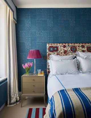 blue bedroom with blue wallpaper, patterned fabric headboard, blue stripe throw and bedding, bedside, pink lamp