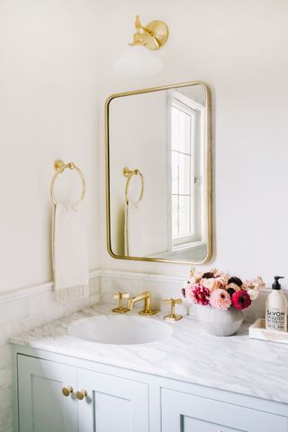 white bathroom with marble topped vanity, pale blue cabinetry, brass fixtures, vase of flowers, wall light, mirror, brass taps
