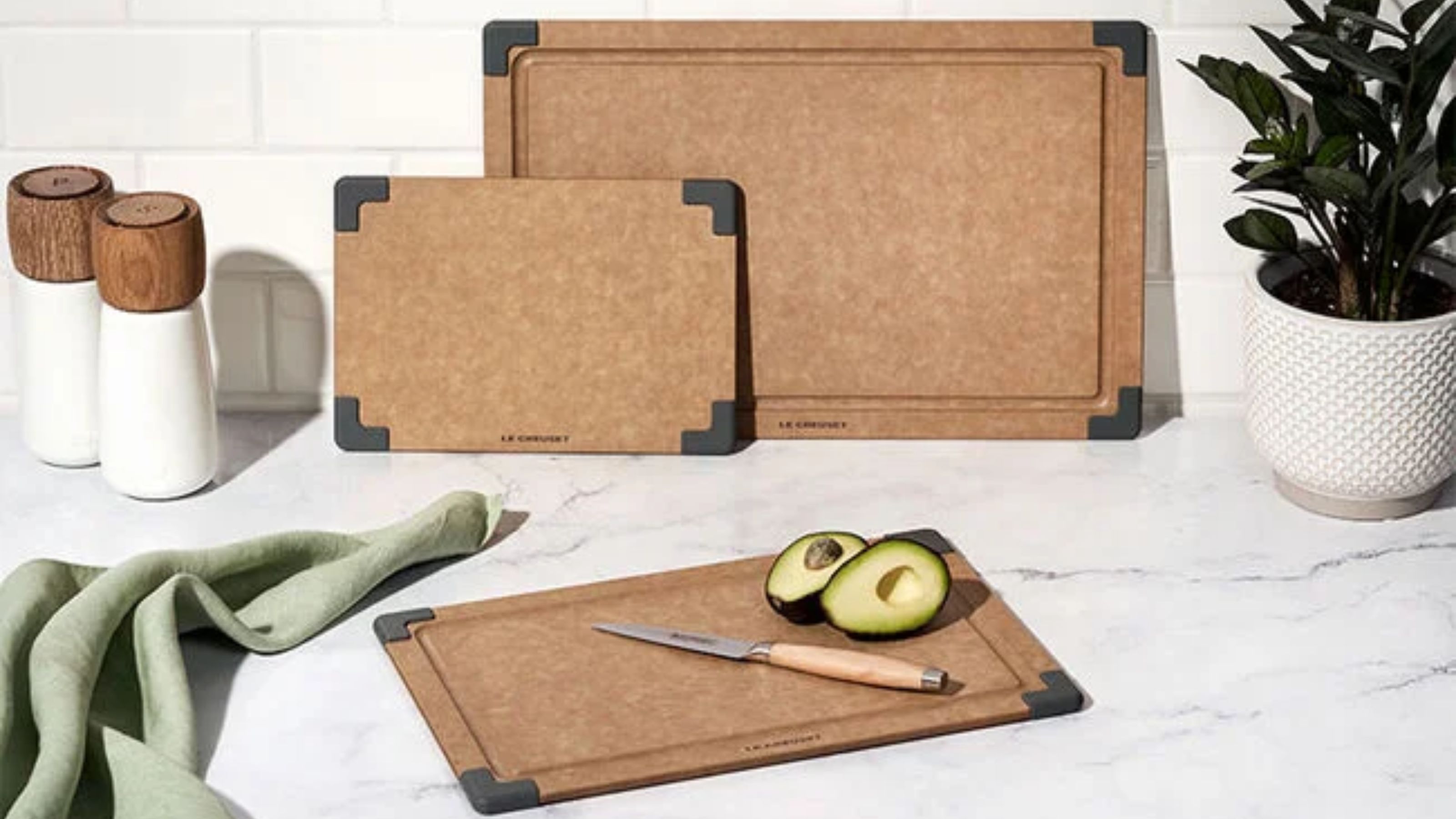 A set of three brown paper fiber cutting boards on a white marble countertop with a white tile backsplash. An avocado is cut in have beside a knife on the smallest of the three.