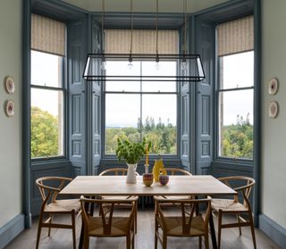 A blue dining room nook painted in Benjamin Moore's Van Courtland blue paint which frames the space beautifully; there is a large pendant light hanging over the wooden dining room table with six wooden chairs around it on top of wood flooring; the window view is of Scottish landscape