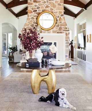 Large entryway with tall brick chimney and fireplace in background, large rounded table with gold sculptural legs, decorated with flowers and ornaments, rug beneath table on wooden flooring, dog sitting in front of table