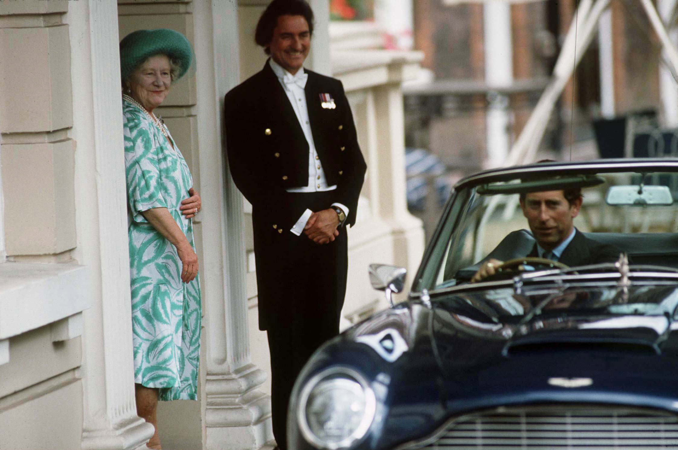 King Charles driving his convertible as the Queen Mother looks on and smiles in a doorway