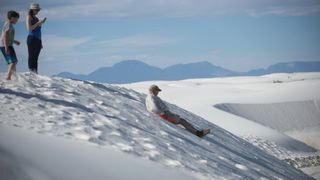 Man sledding down dune at White Sands National Park while two children watch