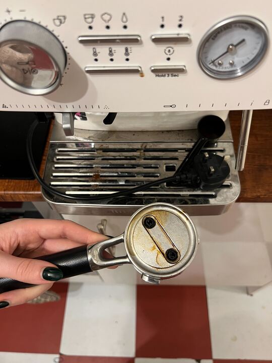 Image of a hand holding a dirty portafiller over red and white tile floors in front of a white Kitchenaid coffee machine.