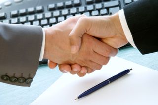 2 men shaking hands over a desk with pen, paper and computer keyboard