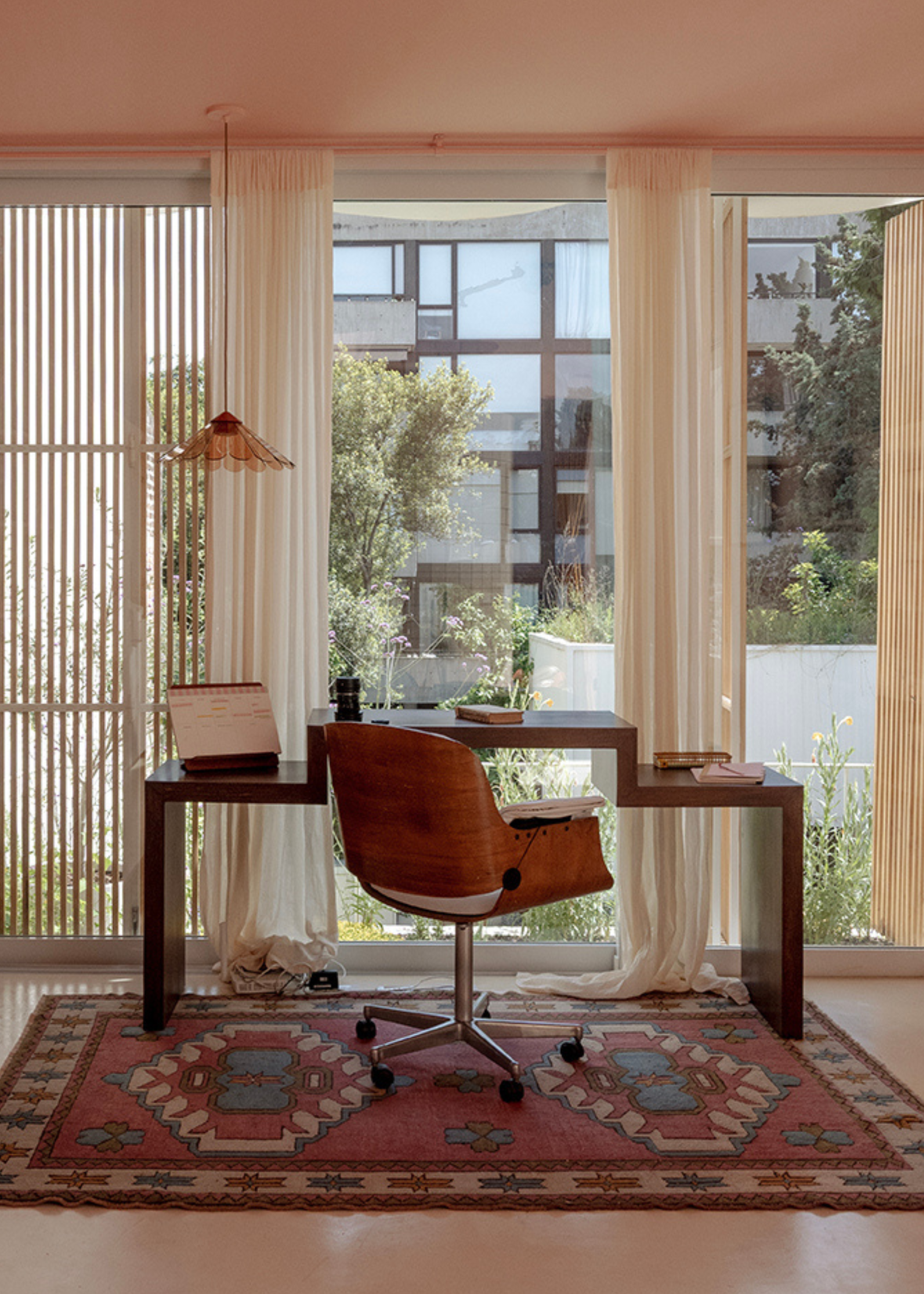 A bright and airy home office with sheer curtains, a wood desk, a swivel chair, and a pendant lamp over a desk calendar