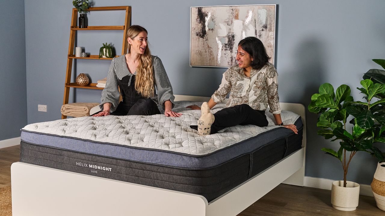 The image shows two mattress testers sat on top of the a white mattress with a navy blue trim. The mattress is placed on a white bed frame in a blue bedroom