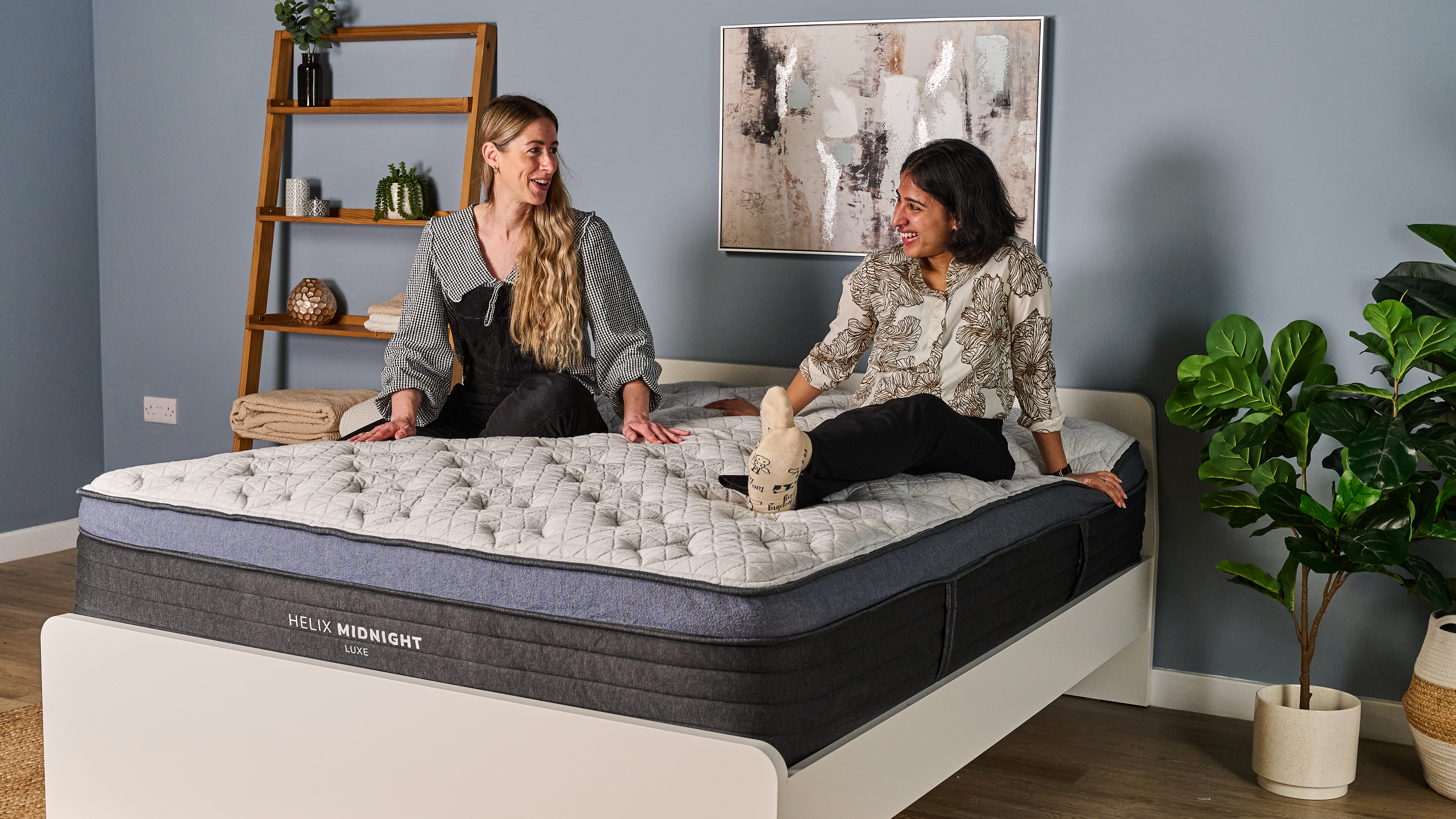 The image shows two mattress testers sat on top of the a white mattress with a navy blue trim. The mattress is placed on a white bed frame in a blue bedroom