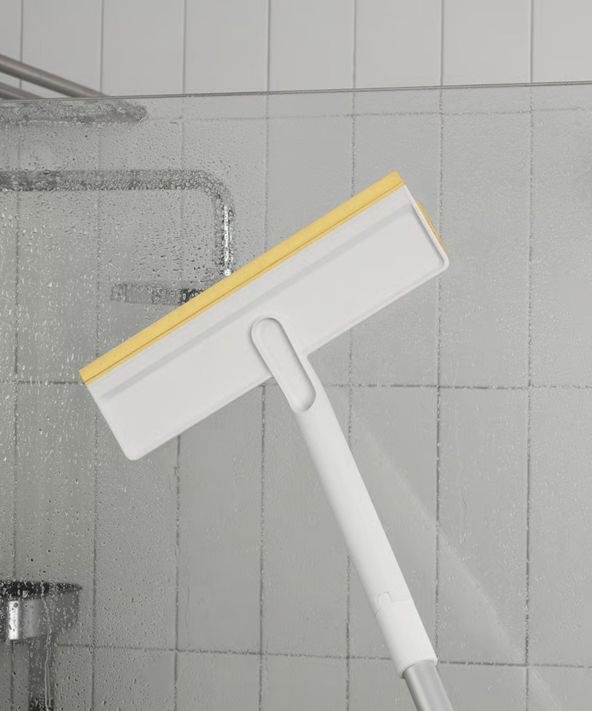 White and yellow IKEA squeegee from the PEPPRIG Cleaning set being used to clean clear shower glass with white tiling and silver pipes shown behind.