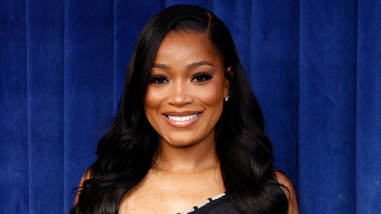 Keke Palmer wearing a black one shouldered dress and smiling against a blue background