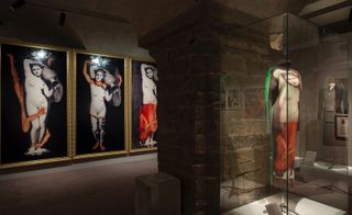 A display of a dress inspired by the human body enclosed in a glass display placed against a concrete wall pillar. Behind the pillar is Yasumasa Morimura’s three-panelled Portrait series hanging on the wall (which inspired the dress)