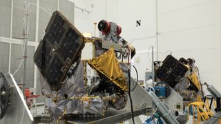 two silver and gold spacecraft sit in a white-walled clean room