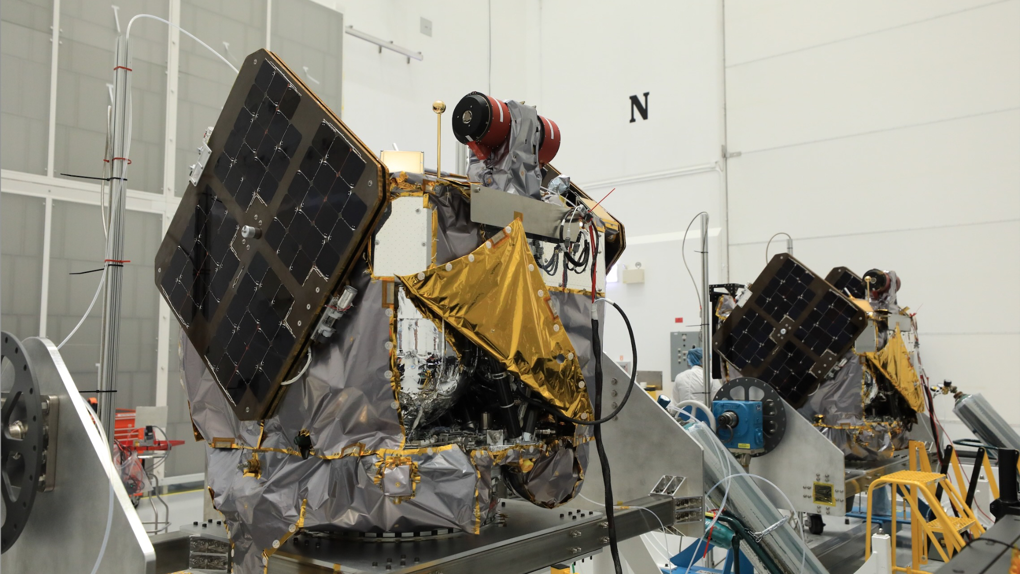 two gold and silver spacecraft sit inside a big, white-walled clean room