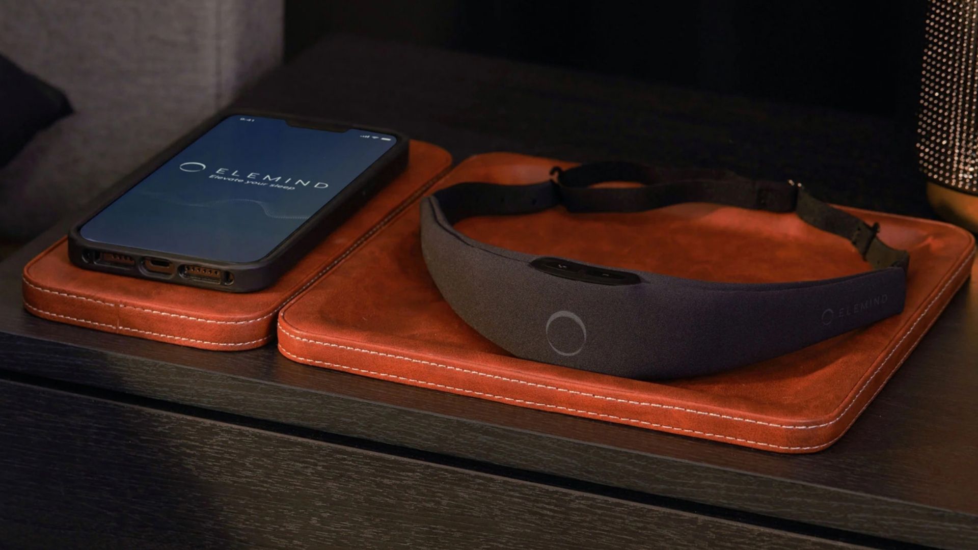 The image shows the black Elemind headband next to a smartphone that's displaying the Elemind app, both placed on a brown leather tray on a nightstand