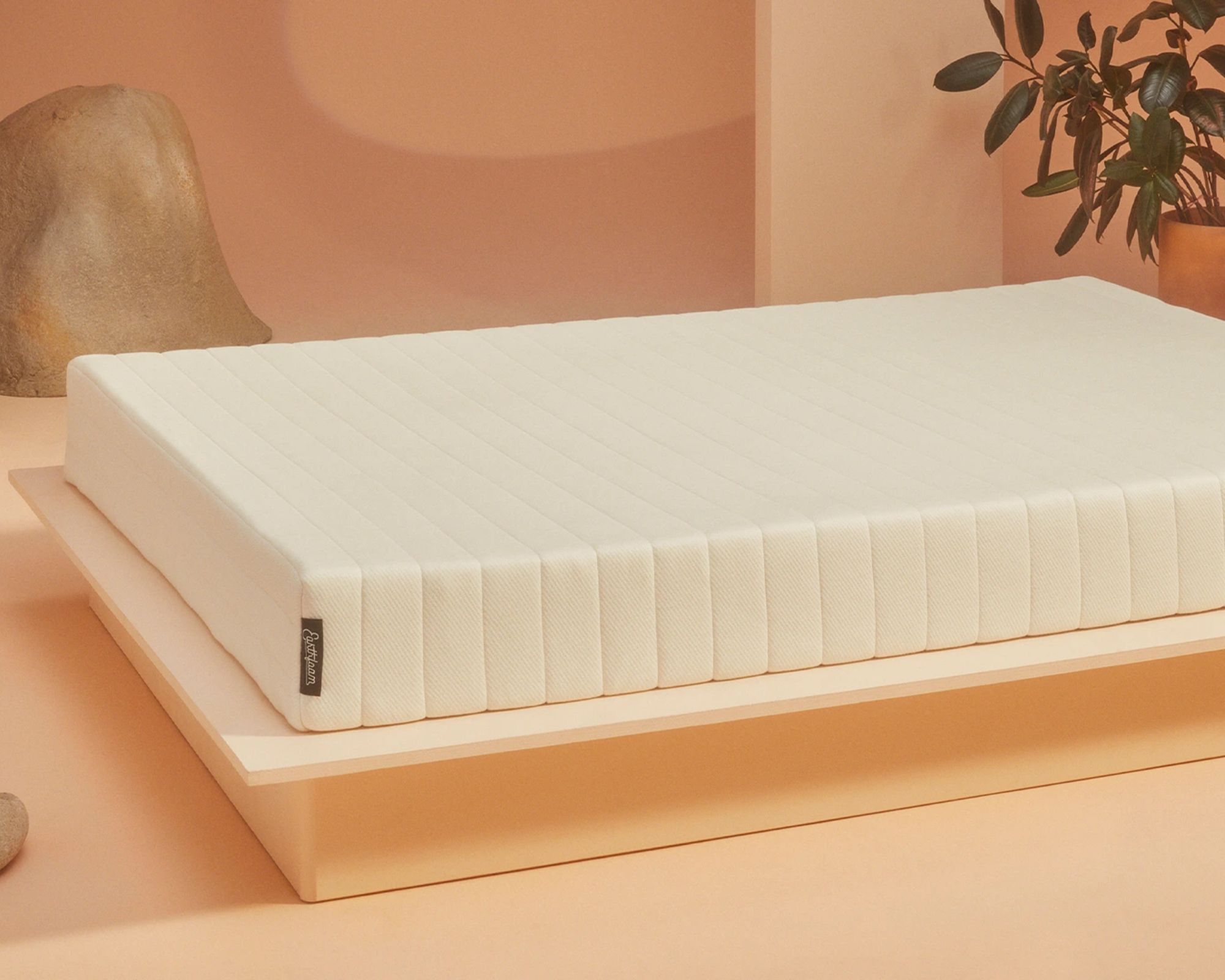 A white Earthfoam organic mattress on a low bedframe in a peach colored room with a houseplant seen behind the mattress in the far right corner.