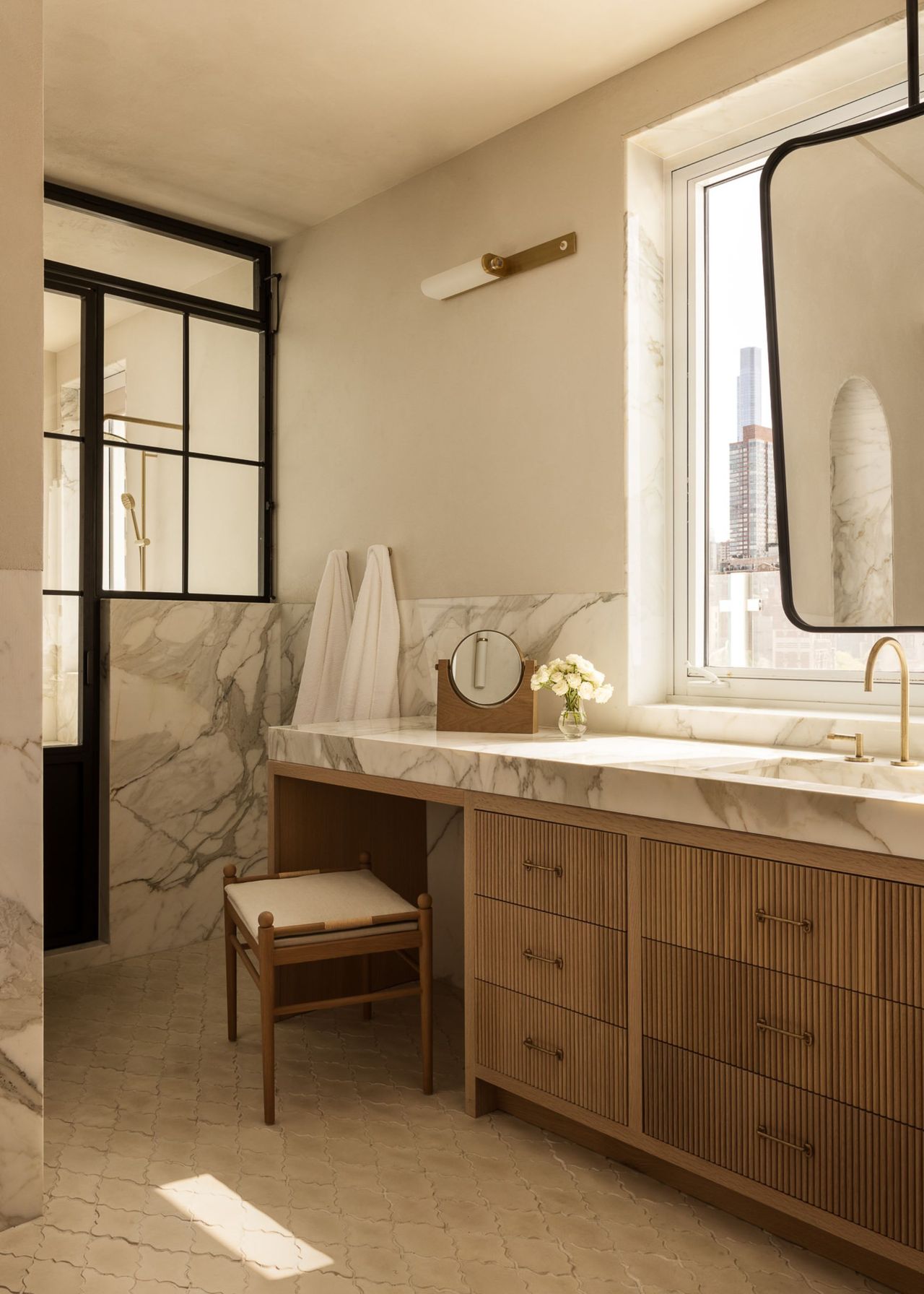 A modern neutral bathroom with wooden drawers with rubbed detailing, a stool by a dressing area section, marble counters and a conservatory style shower