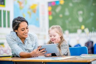 In a classroom, girl with Down syndrome and her teacher look at a tablet computer together. Both are smiling. 