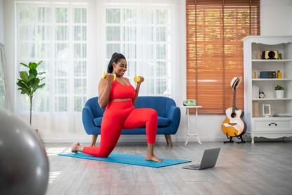 Woman on yoga mat at home lunging with dumbbells curled in arms