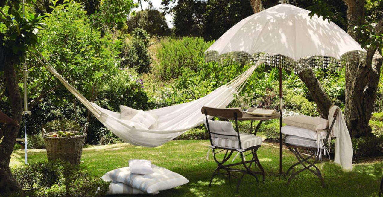 nglish country garden in summer, white cotton hammock, wrought iron and wood garden furniture, white vintage style parasol, floor cushions, large wicker basket full of apples.